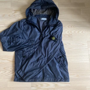 Grå vindjacka från Stone Island - Säljer en grå vindjacka från Stone Island med huva och dragkedja. Tröjan kostar 4700 och är köpt från NK, killen på bilden är typ 170 och den sitter bra. Jackan är i bra skick bara två super små mini jack som ni ser på bild 3 och 4. Hör av er vid minsta fundering 