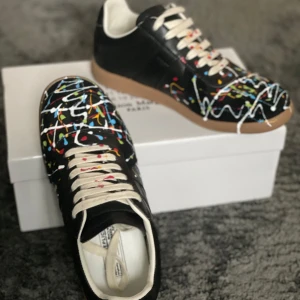 Maison Margiela sneakers med färgstänk 38-39 - Unika svarta sneakers från Maison Margiela med coola färgstänk i vitt, blått, rött, gult och grönt. Skorna har vita snören, beige sula och är tillverkade i skinn. Perfekta för dig som vill sticka ut med en konstnärlig vibe. Använda en gång 
