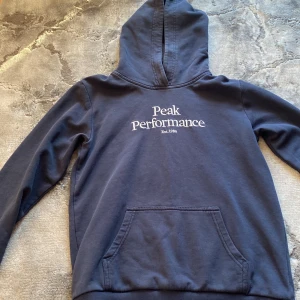 Mörkblå hoodie från Peak Performance - Snygg mörkblå hoodie från Peak Performance med broderad vit logga på bröstet. Klassisk känguruficka framtill, huva och långa ärmar. Tillverkad i mjuk bomull, perfekt för chill dagar eller när du vill ha en clean look.