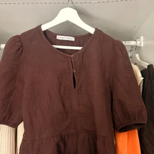 Brun kor blus från Mixray - Supersöt brun kort blus från Mixray i luftig bomull. Klänningen har rund halsringning, knappar framtill och puffiga korta ärmar med resår. Den har en avslappnad passform och volang nedtill för en chill vibe.