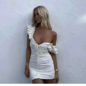 Vit kort klänning med volangdetaljer - Supersöt vit kort klänning med volanger på axel och ärm, snörning framtill och figurnära passform. Perfekt för sommarens alla tillfällen och ger en romantisk vibe. Klänningen har en axelbandslös look på ena sidan och är tillverkad i ett lätt material.