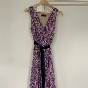 Lång  klänning Carolina Herrera - Säljer en elegant långklänning från Carolina Herrera med lila och beige blommigt mönster. Klänningen har omlott-v-ringad överdel, ärmlös design och ett svart bälte i midjan. Materialet är lätt och luftigt, perfekt för sommarens alla tillfällen.