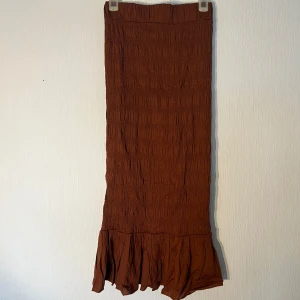 Brun långkjol med volangkant - Snygg brun långkjol med smockad struktur och bred volang längst ner. Kjolen har en rak passform upptill och faller snyggt mot benen. Endast använd få gånger!