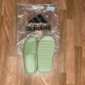 Adidas slides i ljusgrön färg - Adidas slides i en fräsch ljusgrön nyans. De har öppen tå, bred rem över foten och en chunky, räfflad sula för extra grepp. Tillverkade i syntetmaterial som känns mjukt och bekvämt. Perfekta för chill dagar eller efter träningen.