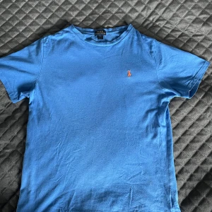 Blå t-shirt från Polo Ralph Lauren - Snygg blå t-shirt från Polo Ralph Lauren med klassisk orange broderad logga på bröstet. T-shirten är i mjuk bomull och har en enkel, stilren design med rund hals och korta ärmar. Perfekt för en chill och avslappnad look.