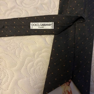 Dolce & Gabbana – Svart slips med diskreta prickar - Elegant och exklusiv slips från Dolce & Gabbana, i sofistikerad svart design med subtila bruna prickar. Ett stilfullt val som lyfter både formella och semi-formella outfits – perfekt för bröllop, affärsmöten eller fest.  Märke: Dolce & Gabbana – Cravatte  Färg: Svart med bruna prickar  Skick: Helt ny, aldrig använd  Förpackning: I original plastförpackning  Tillverkad i: Italien