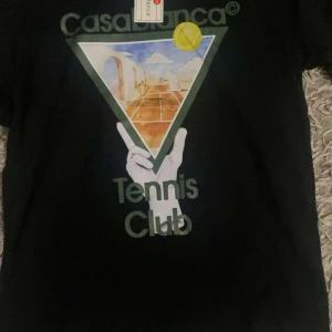 Svart Casablanca Tennis Club t-shirt - Svart t-shirt från Casablanca med stort grafiskt tryck på bröstet som visar en tennisbana, gul tennisboll och texten 'Tennis Club'. Rund hals och korta ärmar. Materialet är mjuk bomull, perfekt för dig som gillar sportig och trendig stil.