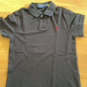 Svart Polo Ralph Lauren t-shirt - Svart t-shirt från Polo Ralph Lauren med klassisk pikékrage och två knappar vid halsen. Slim fit-modell med korta ärmar och röd broderad logga på bröstet. Tillverkad i mjuk bomull som är skön mot huden. Perfekt för dig som gillar stilren och enkel design.