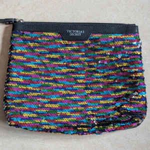 Paljettkuvertväska Victoria's Secret - Färgstark kuvertväska från Victoria's Secret med glittriga paljetter i regnbågens färger på framsidan och svart konstläder på baksidan. Väskan har dragkedja.. Perfekt för dig som vill sticka ut med din stil.
