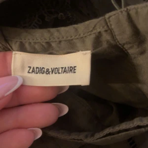 Olivgrön blus från Zadig & Voltaire - Säljer en olivgrön blus från Zadig & Voltaire med hög hals, lång ärm och fina spetsdetaljer vid axlar och nederkant. Blusen har en lös passform och är tillverkad i ett lätt bomullstyg. Perfekt för dig som gillar unika detaljer och stilren design.