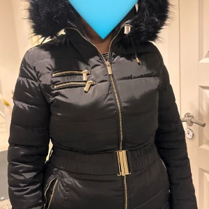 Svart pufferjacka från Roccobarocco - Snygg svart pufferjacka från Roccobarocco med guldfärgade dragkedjor, markerad midja med brett bälte och stor huva med fuskpäls. Jackan har flera fickor med dragkedja och quiltade detaljer. Perfekt för kalla dagar när du vill vara både varm och stilren.