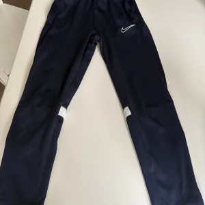 Nike Dri-Fit träningsbyxor marinblå - Snygga marinblå träningsbyxor från Nike med Dri-Fit-teknologi. Byxorna har vita detaljer vid knäna, dragkedjor vid bensluten och elastisk midja. Perfekta för dig som gillar sportig stil och vill ha bekväma byxor till träning eller chill. Liten skråma på baksidan av byxan.