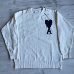 Vit stickad tröja från Ami Paris - Vit stickad tröja från Ami Paris med svart hjärta och bokstaven A broderat på bröstet. Tröjan har rund hals, ribbade muddar och nederkant. Perfekt för dig som gillar stilrena och trendiga plagg med en twist. Storlek S