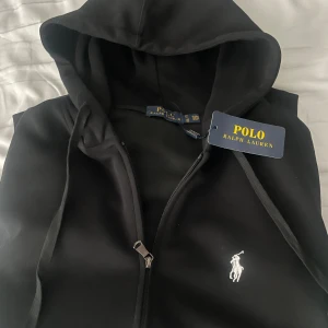 Svart hoodie från Polo Ralph Lauren - Svart hoodie från Polo Ralph Lauren med klassisk huva, dragkedja och snörning. Tröjan har det ikoniska broderade logotypen på bröstet och är tillverkad i mjuk bomull. Perfekt för en chill och stilren look.