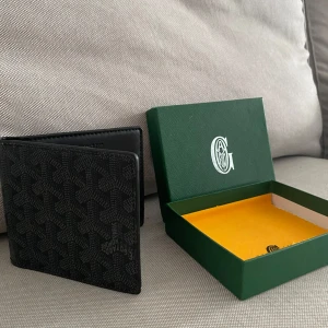 Svart plånbok med mönster - Säljer en svart plånbok från g med deras ikoniska grafiska mönster. Plånboken är i skinn och har en stilren, fyrkantig form. Perfekt för dig som vill ha något exklusivt och snyggt i fickan.