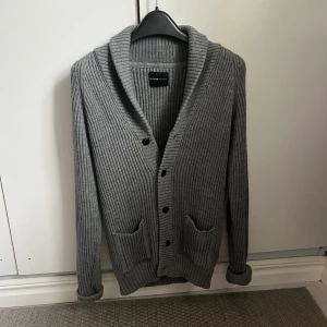 Grå ribbstickad kofta från Selected Homme - Snygg grå ribbstickad kofta från Selected Homme med sjalkrage, knappar framtill och två fickor. Perfekt för lager-på-lager och chillare dagar. Långärmad och i mjukt material som känns skönt mot huden. Tröjan har ingen specifik storlek men sitter som storlek S, Kanske M om man väger lite mindre