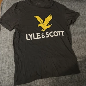 Svart Lyle & Scott t-shirt med tryck - Svart t-shirt från Lyle & Scott med stort gult fågeltryck och vit logga på bröstet. Klassisk passform och rund hals. Perfekt för dig som gillar streetwear och vill sticka ut med en ikonisk logga.