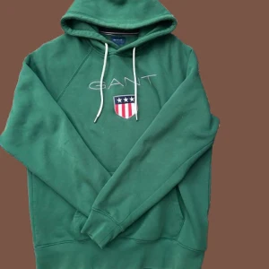 Grön hoodie från GANT med brodyr - Snygg grön hoodie från GANT med vit snörning i huvan och broderad logga samt sköld på bröstet. Klassisk känguruficka framtill och mjukt material som känns skönt mot huden. Perfekt för dig som gillar stilren streetwear.