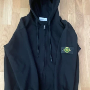 Svart hoodie från Stone Island - Svart hoodie från Stone Island med dragkedja framtill och klassisk logga på ärmen. Tröjan har huva med snörning och ribbade muddar vid ärmslut och nederkant. Perfekt för dig som gillar streetwear och vill ha en stilren look.