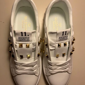 Valentino Rockstud Untitled sneakers - Säljer ett par vita Valentino Rockstud Untitled sneakers med feta guldfärgade nitar och coola detaljer. Skorna har snörning, platt sula och är tillverkade i skinn. Perfekt för dig som vill sticka ut med exklusiv design och edgy vibe.