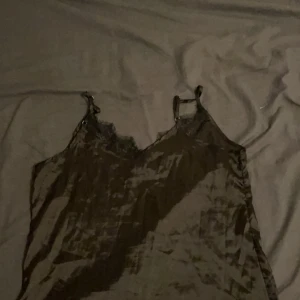 Svart satin linne med spets - Säljer en svart linne i satin med tunna axelband och v-ringning dekorerad med spets. Klänningen har en glansig finish och är perfekt för dig som gillar en stilren och trendig look.