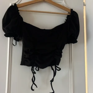 Svart croppad topp med puffärm - Supersnygg svart croppad topp med puffiga korta ärmar och snörning framtill. Toppen har en offshoulder-design och coola rynkade detaljer som ger en trendig look. Perfekt att styla med höga jeans eller kjol för en edgy vibe.