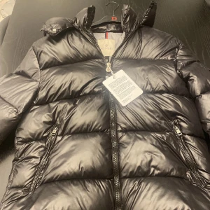 Svart Moncler pufferjacka med huva - Snygg svart pufferjacka från Moncler med glansig finish, huva och två dragkedjefickor framtill. Jackan har en klassisk Moncler-logga på ärmen och quiltad design som ger en riktigt fet vinterlook. Perfekt för dig som vill hålla stilen när det är kallt ute.