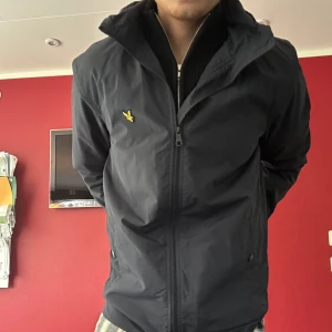 Marinblå vindjacka från Lyle & Scott - Säljer en marinblå vindjacka från Lyle & Scott med klassisk gul logga på bröstet. Jackan har dragkedja framtill, hög krage och två sidofickor. Perfekt för blåsiga dagar och enkel att matcha med olika outfits. Hör av dig vid frågor!😊
