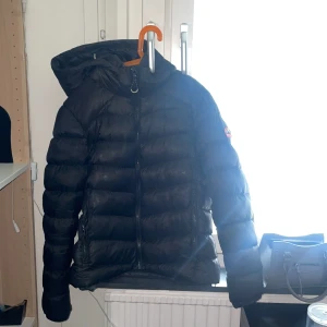 Svart pufferjacka med huva - Säljer en svart pufferjacka från SuperDry med stor huva och dragkedja framtill. Jackan har ett broderat logomärke på ärmen och är quiltad för extra värme. Perfekt för kalla dagar och är figursydd. Den är lite smutisg vid luvan se bild