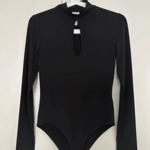 Svart långärmad bodysuit med cut out - Säljer en svart långärmad bodysuit med hög hals och snygg cut out-detalj framtill. Tight passform som sitter snyggt och ger en clean look. Perfekt att styla med jeans eller kjol för en trendig outfit