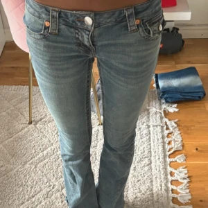 True Religion Joey Low Rise Flare Jeans - Säljer ett par ljusblå True Religion Joey jeans med låg midja och flare-ben. Klassiska femficksmodellen med snygga detaljer på bakfickorna och silverfärgade knappar. Jeansen är använda en gång och i nyskick. Färgen säljs inte längre.