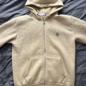 Moncler Hoodie - Säljer en beige hoodie från Moncler med huva och hel dragkedja framtill. Tröjan har två fickor och Moncler-logga broderad på bröstet. Perfekt för chill dagar och enkel att matcha med jeans eller joggers.