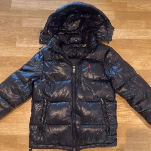 Svart dunjacka Polo Ralph Lauren - Snygg svart dunjacka från Polo Ralph Lauren med glansig finish, röd broderad logga på bröstet och avtagbar huva. Jackan har dragkedja framtill och två praktiska fickor med dragkedja. Perfekt för kalla dagar och riktigt stilren look.