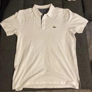 Vit Lacoste pikétröja i bomull - Vit pikétröja från Lacoste med klassisk krage och knappslå. Ikoniska gröna krokodilloggan broderad på bröstet. Tröjan är kortärmad och tillverkad i mjuk bomull, perfekt för en clean och chill stil. Passformen är normal och materialet känns riktigt skönt mot huden. Helt oanvänd