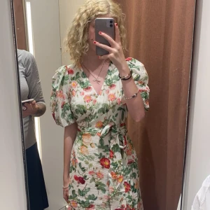 Blommig omlottklänning med puffärm - Superfin blommig omlottklänning från Gina Tricot med v-ringning, puffiga korta ärmar och knytband i midjan. Klänningen är vit i grunden med färgglada blommor i rött, rosa och grönt. Perfekt för sommarens alla tillfällen!