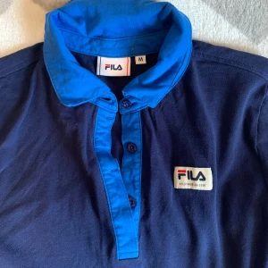 Blå pikétröja från FILA med snörning - Snygg marinblå pikétröja från FILA med kontrasterande blå krage och knappslå. Tröjan har klassisk pikédesign, broderad FILA-logga på bröstet och praktisk snörning nertill för en cool detalj. Perfekt för dig som gillar sportig och avslappnad stil.