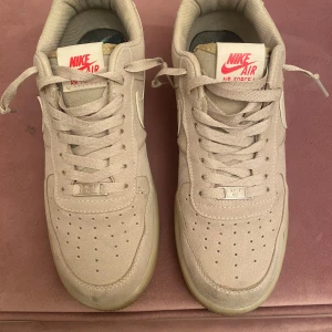 Nike Air Force 1 i beige mocka - Snygga Nike Air Force 1 sneakers i beige mocka med klassisk vit swoosh och röd Nike-logga på plösen och hälen. Lågt skaft, snörning och perforerad tå för extra ventilation. Perfekta för dig som gillar stilrena och tidlösa sneakers med en twist.