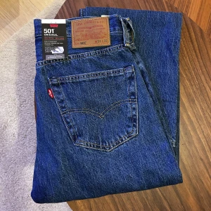 Levi's 501 blå jeans W31 L32 (helt nya) - Klassiska Levi's 501 jeans i mörkblå denim. Rak passform med normal midja och ikonisk femficksdesign. Tillverkade i 100% bomull för en autentisk känsla. Perfekta för dig som gillar tidlös stil och vill ha ett par riktigt schyssta jeans.