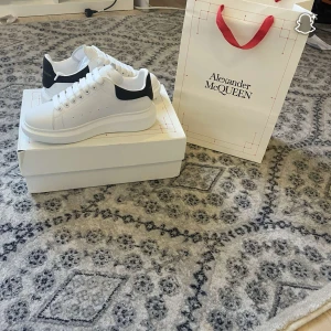 Alexander McQueen vita sneakers - Snygga vita sneakers från Alexander McQueen med svart häl och guldfärgad logga. Skorna har chunky plattformssula, rund tå och breda vita skosnören. Tillverkade i skinn för en lyxig känsla och stilren look. Perfekta för dig som vill sticka ut med klass.