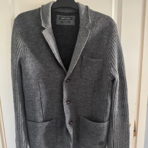 Marc O'Polo cardigan - Snygg grå kavaj från Marc O'Polo i kokt ull med stickade ärmar och tre bruna knappar framtill. Kavajen har tre praktiska fickor och en klassisk krage med råa kanter. Perfekt för dig som gillar en chill men stilren look.