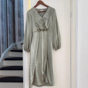 Ljusgrön långklänning från Bubbleroom - Snygg ljusgrön långklänning från Bubbleroom med glansig finish och v-ringad omlott upptill. Klänningen har långa ärmar med resår vid handlederna och är gjord i ett satinliknande material som ger en lyxig känsla. Perfekt för dig som vill sticka ut med stil.