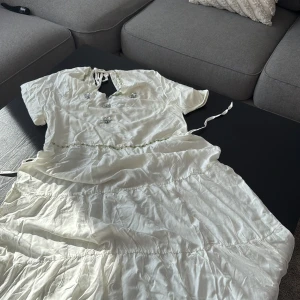 Vit långklänning med pärldetaljer - Superfin vit långklänning från H&M med kort ärm, knytning i nacken och gröna pärldetaljer längs midjan och halsringningen. Klänningen har dekorativa silverblommor framtill och ett flowigt, romantiskt fall. Perfekt för dig som vill sticka ut med en unik look. Loop