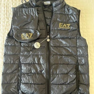 Svart dunväst från Emporio Armani EA7 - Snygg svart dunväst från Emporio Armani EA7 med guldfärgad logga på bröstet. Västen är quiltad, har dragkedja framtill och levereras med tillhörande förvaringspåse. Perfekt för lager-på-lager och enkel att ta med sig.