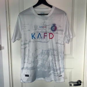 Vit Al Nassr Ronaldo 7 fotbollströja - Snygg vit fotbollströja från Al Nassr med Ronaldo och nummer 7 på ryggen. Tröjan har korta ärmar, tryckta loggor och sponsortryck i blått och rött framtill. Materialet är lätt och andas, perfekt för träning eller match. Unik design med subtilt mönster över hela tröjan. 