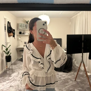 Vit blus med volanger och svarta detaljer från Neo Noir - Säljer en superfin vit blus med volanger och svarta dekorband. Blusen har v-ringad hals, omlottkänsla och långa ärmar med volangdetaljer. Materialet är lätt och luftigt, perfekt för vår och sommar. Passar dig som gillar romantisk och trendig stil. Nästan aldrig använd 