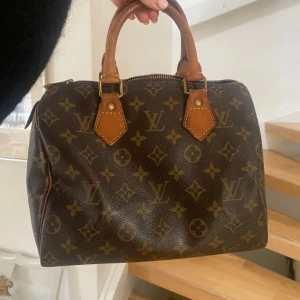 Louis Vuitton monogram handväska - Säljer en klassisk Louis Vuitton Speedy handväska i brunt skinn med ikoniskt monogrammönster och detaljer i ljusbrunt läder. Väskan har två handtag och dragkedja upptill. Perfekt storlek för vardagens essentials och en riktig statement piece. Fint skick men några slitage (se sista bilden ). Äkthetskontrollerad av vestiare collective 