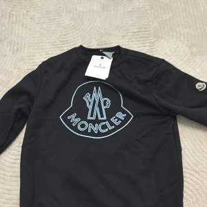 Svart Moncler sweatshirt med logga - Svart sweatshirt från Moncler med stor ljusblå logga på bröstet och klassisk rund hals. Tröjan har långa ärmar och en Moncler-patch på vänster ärm. Perfekt för dig som gillar exklusiva streetwear-vibbar och tydliga designer.