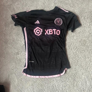 Inter Miami Messi 10 fotbollströja XS - Snygg svart Inter Miami fotbollströja från Adidas med rosa detaljer och tryck. Messi 10 på ryggen, klubbmärke på bröstet och sponsorlogga framtill. Slim fit-modell i lätt och ventilerande material, perfekt för match eller träning.