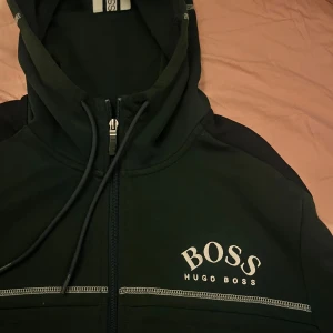 Svart hoodie från Hugo Boss med dragkedja - Säljer en svart hoodie från Hugo Boss med vit logga på bröstet. Tröjan har hel dragkedja, huva med snörning och snygga kontrastsömmar. Köpt i London, finns lite tecken på slitning annars perfekt. Tröjan tillverkas ej längre och är sällsynt.
