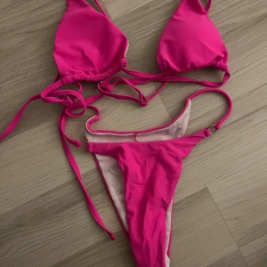 Rosa bikini med knyt och ringdetaljer - Säljer en snygg trekantsbikini i stark rosa från Yigelisa. Bikinin har smala band med knyt i sidorna och små ringdetaljer på både över- och underdel. Materialet är mjukt och stretchigt i polyester och elastan, perfekt för stranden eller poolen.